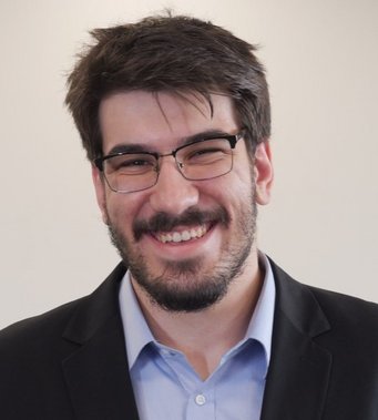 Person with short, dark hair, dressed in a black suit jacket over a light blue collared shirt, standing against a neutral background.