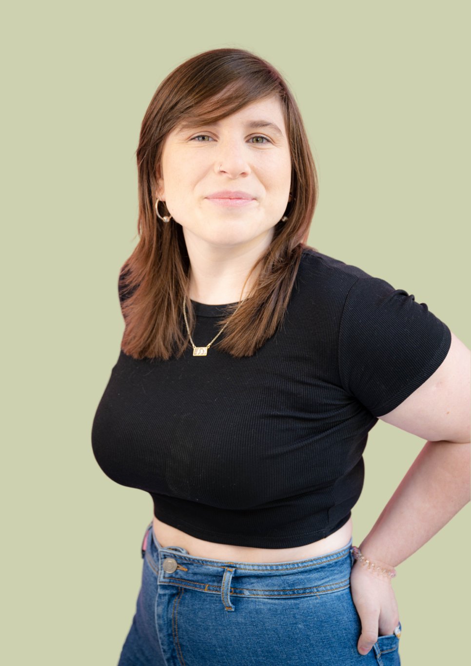 Woman dressed casually in a black ribbed shirt and blue jeans, standing with a hand on her hip. She wears a necklace and bracelet.