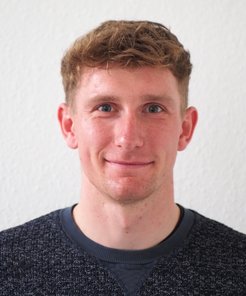 A person with short, curly brown hair and a dark textured sweater stands in front of a white wall, highlighting the attire.