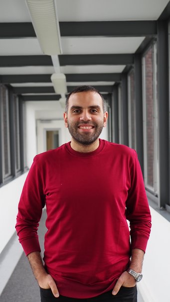 A person wearing a red sweater and a silver wristwatch stands in a modern corridor with large windows and gray flooring.