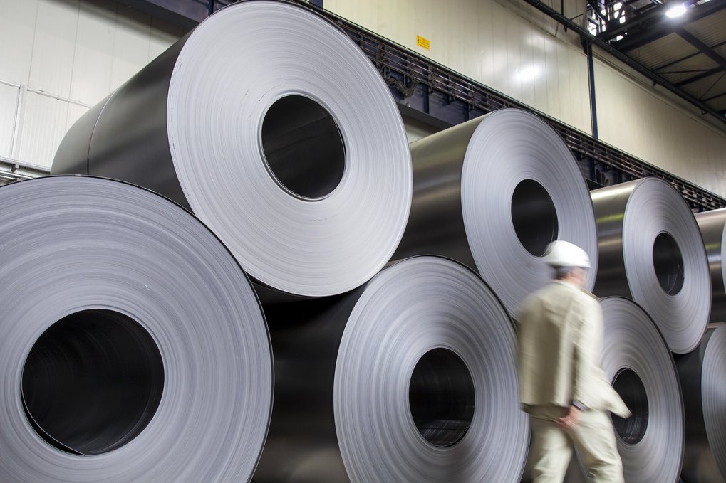 Ein Mann in hellem Anzug und mit weißem Schutzhelm läuft an mannshohen Rollen von Stahlblechen vorbei, die in zwei Reihen übereinander gestapelt sind.
