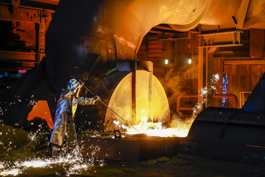 Stahl aus der Ökoschmiede: In einer Pilotanlage in Duisburg ersetzt ThyssenKrupp bei der Eisenverhüttung einen Teil der Kohle durch Wasserstoff und verringert den CO2-Ausstoß auf diese Weise um bis zu 19 Prozent. Eine Person in einem metallisch glänzenden Hitzeschutzanzug steckt ein Gerät mit langer Stange in eine Wanne mit geschmolzenem Eisen. Im Hintergrund ist ein zylinderförmiger Kessel zu sehen.