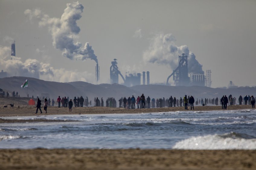 Industrie vor dem Umbruch: Stahlproduktion wie etwa im Stahl- und Hüttenwerk Tata Steel im niederländischen Ijmuiden ist für Industriegesellschaften unverzichtbar. Doch um die Klimaziele zu erreichen, muss sie dafür künftig vermehrt Energie aus regenerativen Quellen nutzen. Im Vordergrund ein Gewässer, an dessen sandigem Ufer zahlreiche Menschen stehen oder gehen. Im Hintergrund leicht von Dunst eingehüllt ein Stahlwerk mit den Hochöfen und rauchenden Kaminen.
