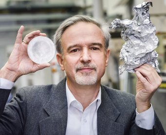 Vordenker einer nachhaltigen Metallindustrie: Dierk Raabe sieht unter anderem im vermehrten Recycling etwa von Aluminiumschrott eine Möglichkeit, den CO2-Fußabdruck der Branche zu reduzieren. frontales Porträt von Dierk Raabe: graumelierte Haare, kurzer Schnurr- und Kinnbart, weißes Hemd und graues Nadelstreifen-Kacket. Er hält in der linken Hand ein Stück zerkitterte Alufolie und in der rechten ein rundes Stuck Aluminium.