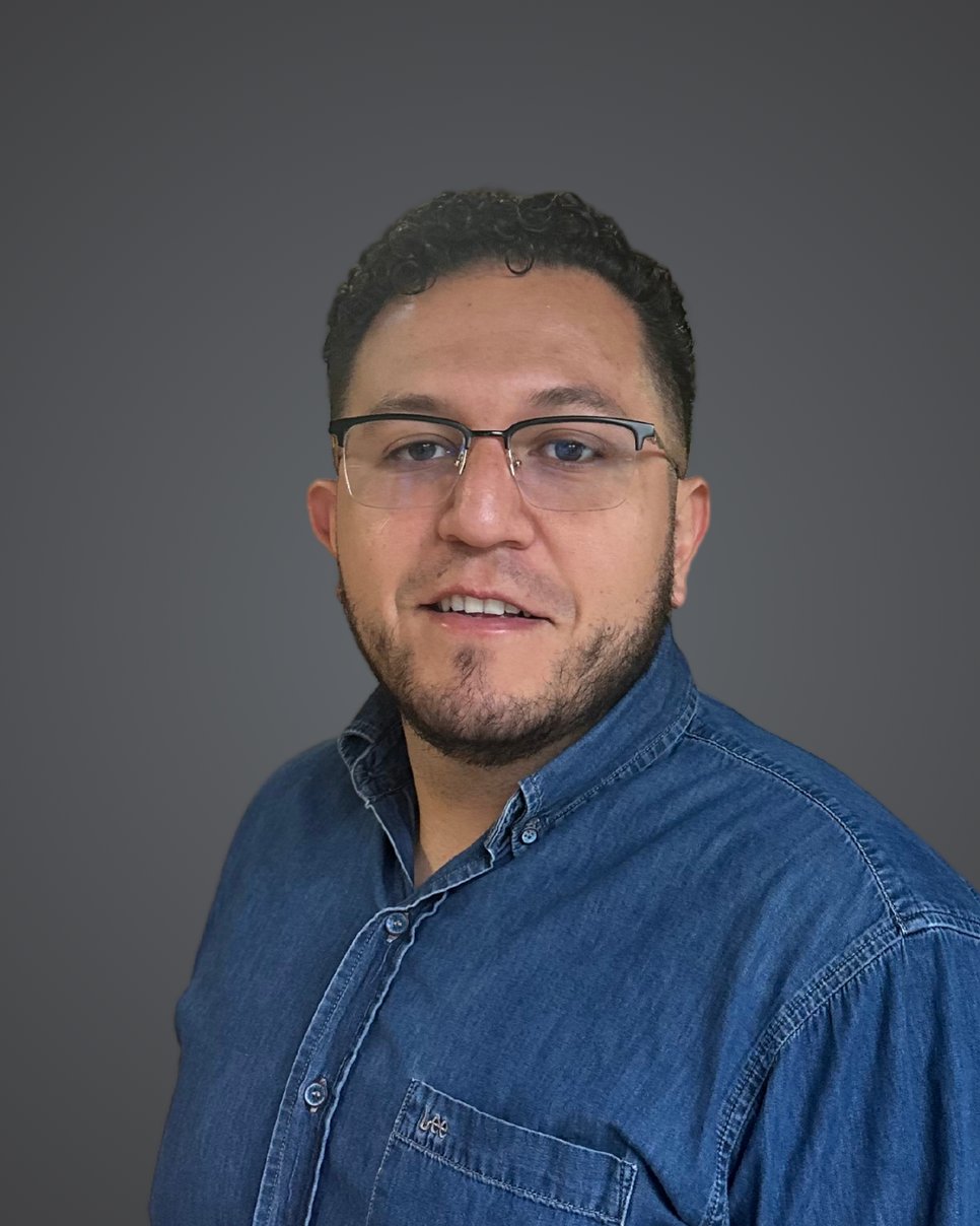 A person with curly hair, dressed in a dark blue button-up denim shirt, against a plain gray background.