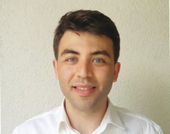 Person dressed in a crisp white shirt, posed in front of a neutral beige background. Hair is neatly styled, with short sideburns visible.