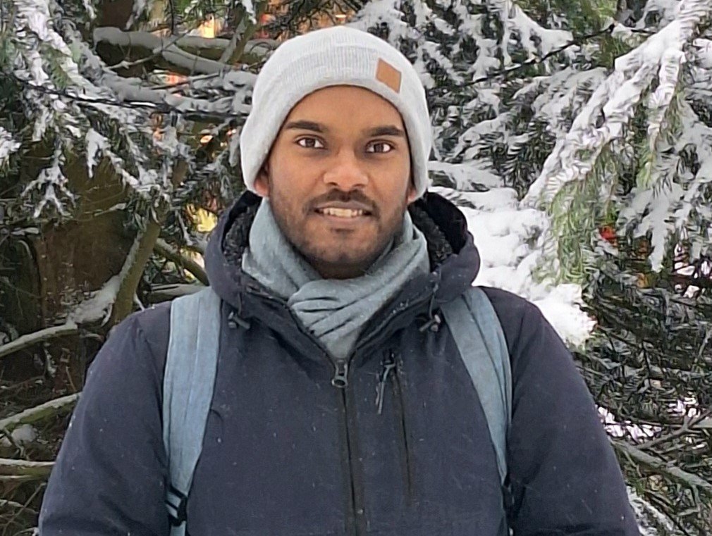 A person in a dark winter jacket, light scarf, and white beanie stands outdoors, surrounded by snow-covered evergreen branches. A backpack is visible.