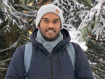 A person in a dark winter jacket, light scarf, and white beanie stands outdoors, surrounded by snow-covered evergreen branches. A backpack is visible.