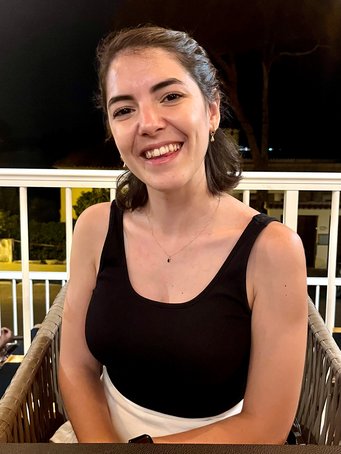 A woman wearing a sleeveless black top and delicate necklace sits in a wicker chair on a terrace at night, with trees and buildings visible in the background.