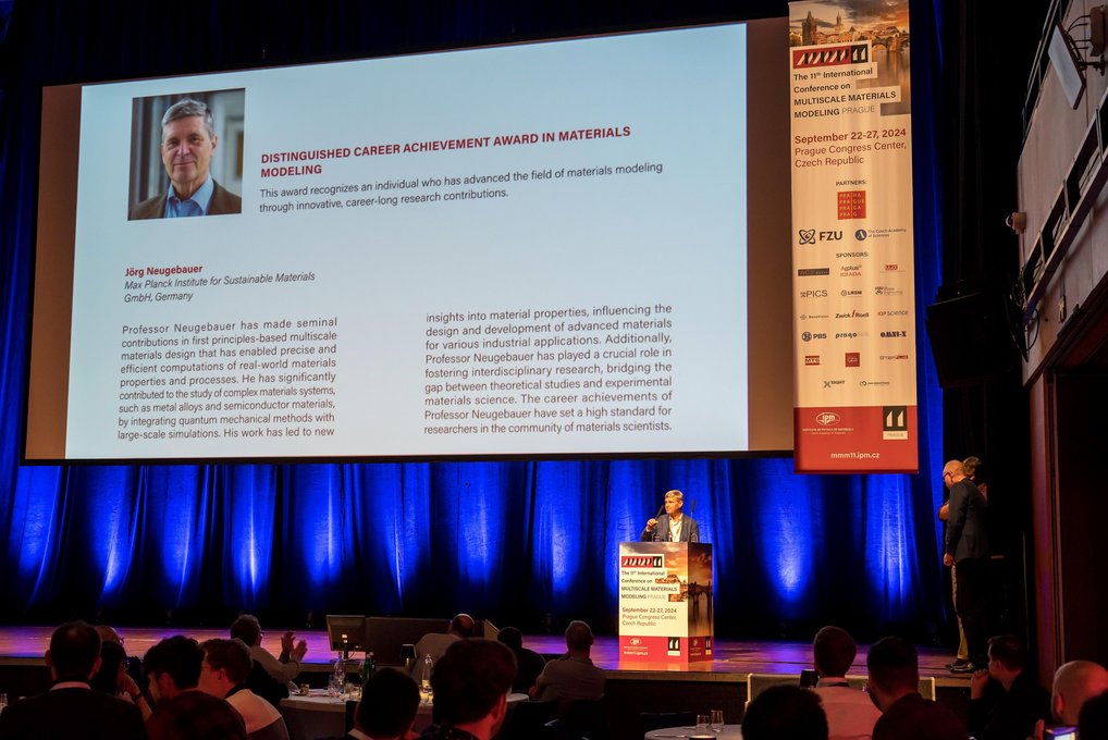Professor Jörg Neugebauer giving the Award Lecture during the 11th International Conference on Multiscale Materials Modeling. Presentation of an award at a conference with a large screen and speaker.