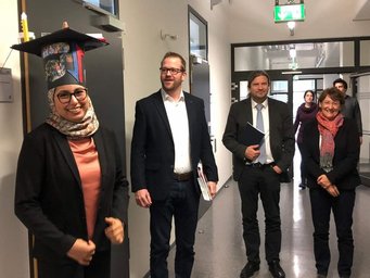 In a modern hallway, five people are gathered, some holding documents, one wearing a graduation cap adorned with vibrant decorations.