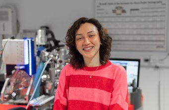 Wie lassen sich Permanentmagnete ohne Seltene Erden herstellen ohne an Leistungsstärke einzubußen? Dies erforscht Larissa Feitosa am MPI-SusMat. Person in rotem Pullover vor Laborgeräten und Computermonitoren.