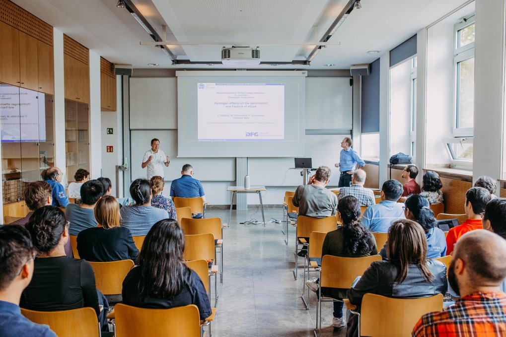 Scientific Curriculum A group of people seated in a university lecture hall attentively follow a presentation on the effects of hydrogen adsorption, presented by two speakers with a projected slide detailing research supported by the DFG.