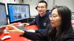 Zwei Personen sitzen an einem Tisch mit roter Oberfläche und betrachten wissenschaftliche Bilder auf einem Monitor. Eine Person zeigt auf den Bildschirm, während die andere die Tastatur bedient. Im Hintergrund sind technische Geräte zu sehen.