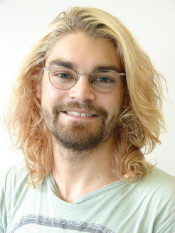 Shoulder-length, curly, blond hair, worn with a light green T-shirt showing a gray stripe.