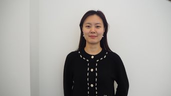 A person stands facing forward, wearing a black sweater with decorative white stitching and buttons. The background is a plain white wall.