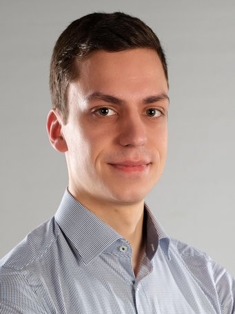 Individual with short brown hair, dressed in a formal blue-striped shirt. The setting features a neutral gray background.