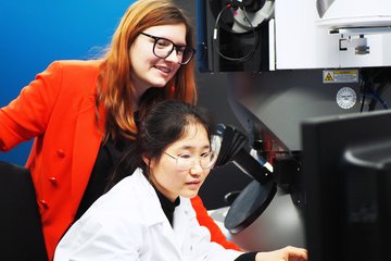 In a laboratory, two individuals focus on a microscope; one wears a white lab coat, the other one, Dr. Lilian Vogl, in a bright red blazer.