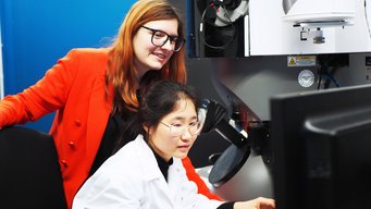 Dr. Lilian Vogl (left) is heading the new group “Environmental & Analytical Electron Microscopy”. The group focuses on advancing materials for green technologies. In a laboratory, two individuals focus on a microscope; one wears a white lab coat, the other one, Dr. Lilian Vogl, in a bright red blazer.