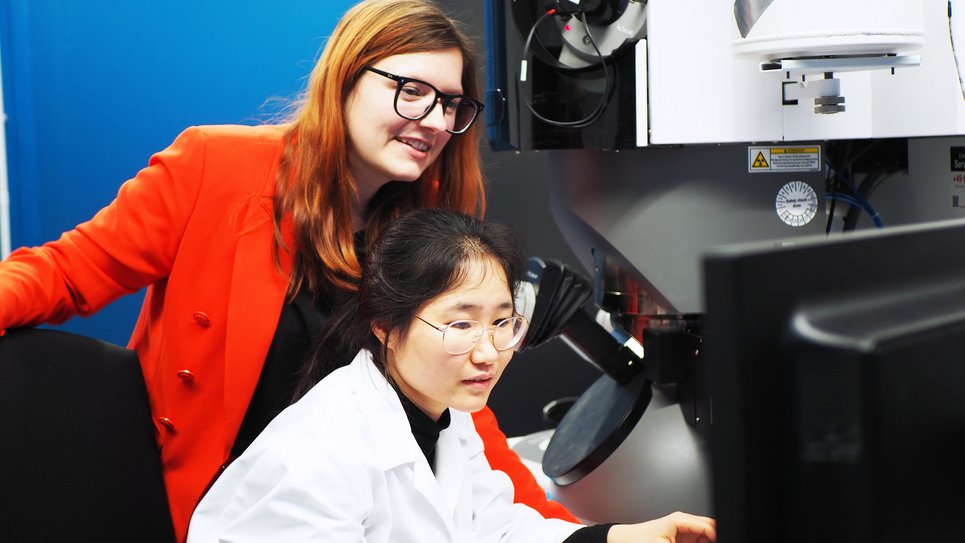 In a laboratory, two individuals focus on a microscope; one wears a white lab coat, the other one, Dr. Lilian Vogl, in a bright red blazer.