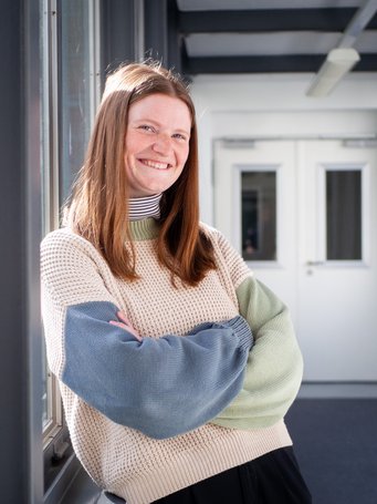Person in a brightly lit hallway, sporting a beige knitted sweater with blue and green sleeves. Sunlight filters through the window.