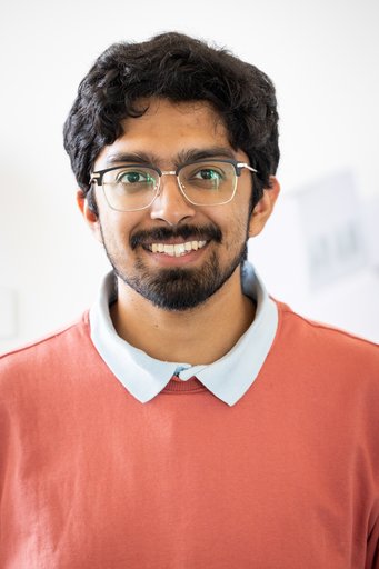 Person with dark curly hair dressed in a red top over a white collared shirt, standing against a light background.