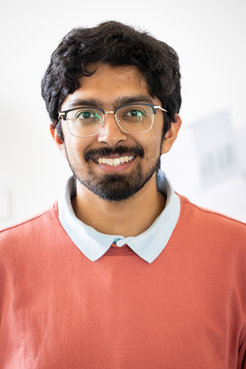 Person with dark curly hair dressed in a red top over a white collared shirt, standing against a light background.