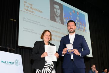 Zwei Personen auf einer Bühne; eine hält eine Medaille, die andere eine Urkunde. Im Hintergrund eine Präsentationsfolie mit Informationen über eine Dissertation im Bereich Materialphysik und Chemie. Ein Logo der Max-Planck-Gesellschaft ist sichtbar.