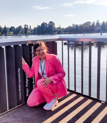 Dr. Lilian Vogl beim Nobelpreisträger-Steg, wo die Namen und Errungenschaften der NobelpreisträgerInnen aufgelistet sind. Person in rosa Anzug kniet auf Pier bei sonnigem Wetter.