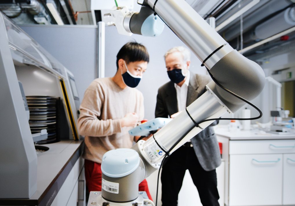 Robotic arm in a laboratory, two individuals collaborating in the background.