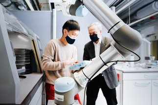 Robotic arm in a laboratory, two individuals collaborating in the background.