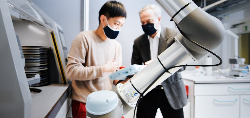 Robotic arm in a laboratory, two individuals collaborating in the background.