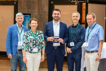 Dr. Tim M. Schwarz (Mitte) mit Prof. Jörg Neugebauer, Prof. Christina Scheu, Prof. Dierk Raabe und Prof. Gerhard Dehm (von links nach rechts), Direktoren und Abteilungsleiterin des MPI-SusMat, nach der Preisverleihung in Brandenburg, Deutschland. Fünf Personen mit Konferenz-Badges, mittlere Person hält eine Medaille.