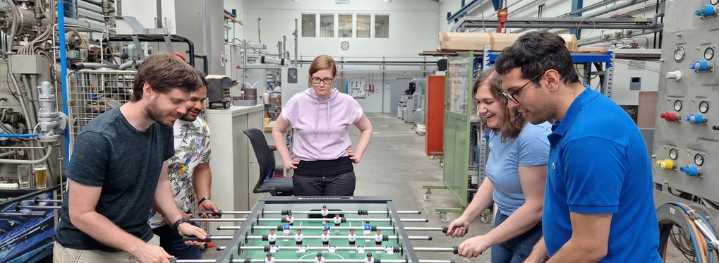 In an industrial setting, four individuals are playing table football, surrounded by machinery and equipment, under a high ceiling with skylights.
