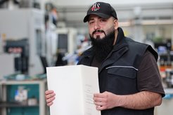 Person in black vest and cap holding a document. Industrial machinery can be seen in the background.