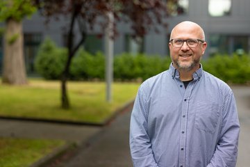 Person mit Brille steht vor einem Gebäude mit Bäumen im Hintergrund.