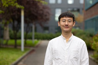 The German Research Foundation (DGM) awarded Dr. Liuliu Han with the DGM Prize for Young Scientists. Han is project group leader at the Max Planck Institute for Sustainable Materials and focusses on developing rare-earth free magnets. Person in white shirt standing outdoors near building. Trees are seen in the background.