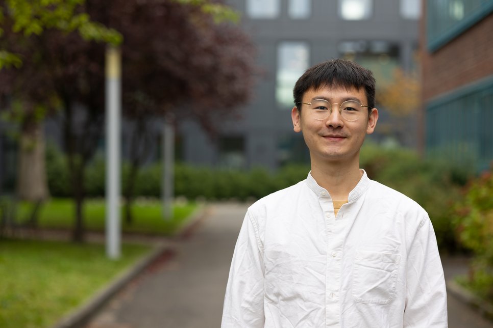 Person in white shirt standing outdoors near building. Trees are seen in the background.