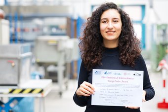 Yosra Dammak, Doktorandin am MPI-SusMat, gewann den Young Poster Prize auf der 16. International Conference on Advanced Lithium Batteries for Automotive Applications. Ihre Arbeit konzentriert sich auf die Verbesserung von Kathodenmaterialien für Lithium-Ionen-Batterien. Eine Person mit lockigem Haar hält ein Zertifikat. Im Hintergrund ist eine wissenschaftliche Laborumgebung.