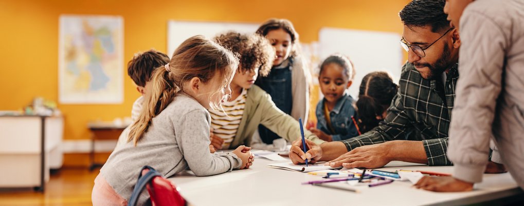 Kinder diskutieren und zeichnen an einem Tisch, umgeben von bunten Stiften und Papier. Im Hintergrund eine Weltkarte an der Wand.