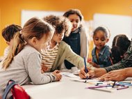 Kinder diskutieren und zeichnen an einem Tisch, umgeben von bunten Stiften und Papier. Im Hintergrund eine Weltkarte an der Wand.