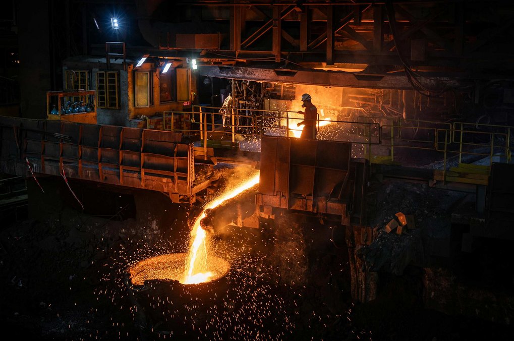 In a dark hall, brightly glowing metal flows from a furnace into a round, man-sized tub. A man wearing a hard hat stands in front of the furnace.