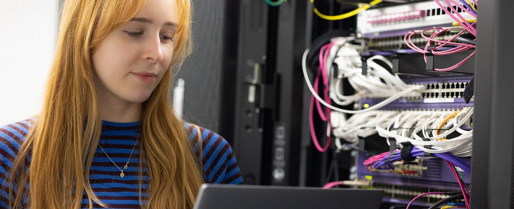 Frau schaut auf Laptop. Neben ihr im Hintergrund ist sind Server mit bunten Kabeln.