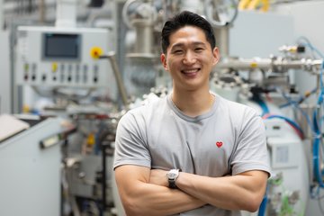 Man in a gray t-shirt with arms crossed standing in front of industrial machinery.