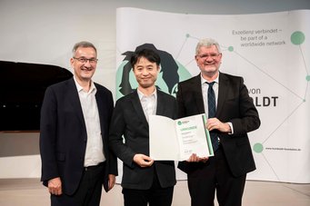 Prof. Sang Ho Oh (Mitte) nahm persönlich seine Urkunde am 24. Juni 2026 bei der Humboldt-Jahresversammlung entgegen.Links neben ihm steht Peter Kreisler (Bundesministerium für Forschung, Technologie und Raumfahrt; Leiter der Unterabteilung "Hochschulen"), rechts Robert Schlögl, Präsident der Humboldt-Stiftung. Drei Männer in schwarzen Anzügen. Zwei von ihnen halten gemeinsam eine Urkunde.