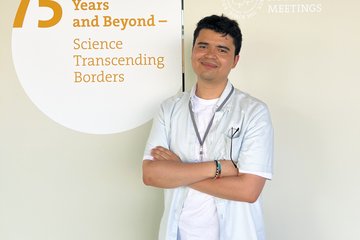 Man standing in front of a beige wall with a text about the 75 anniversary of the Lindau Nobel Laureate Meeting.