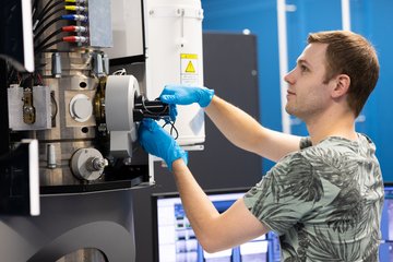 Man wearing blue gloves operating a high-tech laboratory device with multiple cables and control panels.
