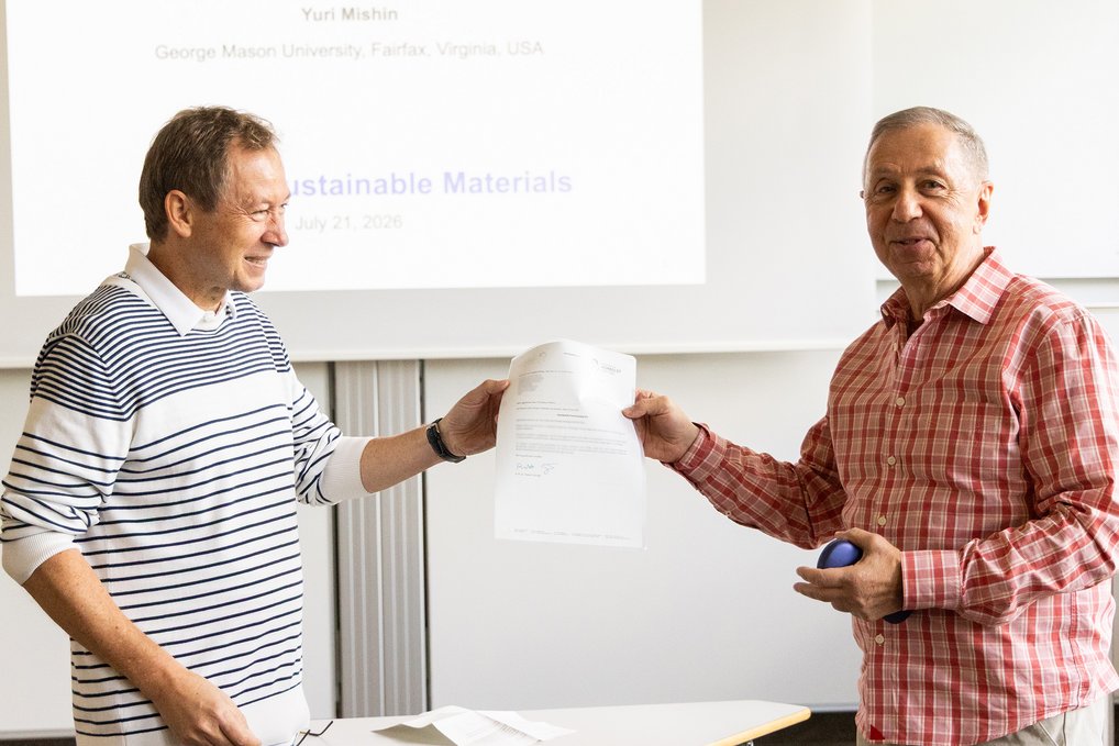 A man hands over a certificate to another man. Both are standing in a seminar room.
