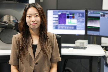 Woman sitting in laboratory. Behind her are monitors showing scientific results.