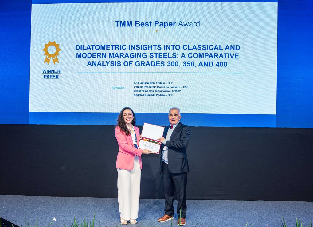 Man hands over a certificate to a woman. They are standing on a stage. A presentation is shown in the background.